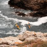 Two kayakers paddle through waves near rocky cliffs on a vibrant summer day.