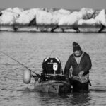 Monochrome image of a man fishing from a kayak near a rocky shore, conveying a serene outdoor atmosphere.