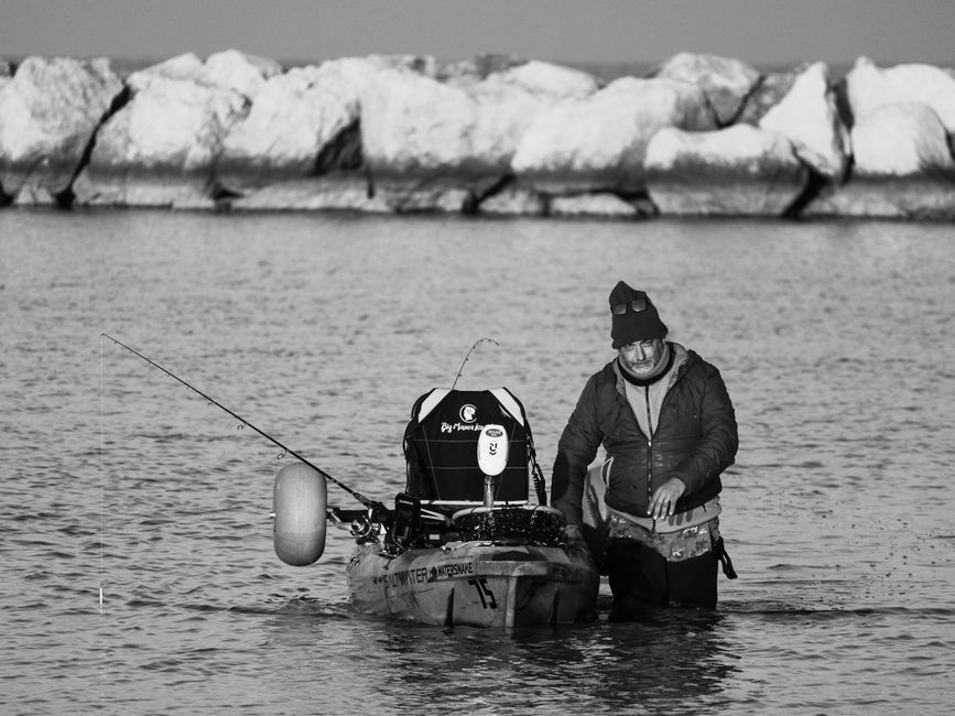 Monochrome image of a man fishing from a kayak near a rocky shore, conveying a serene outdoor atmosphere.
